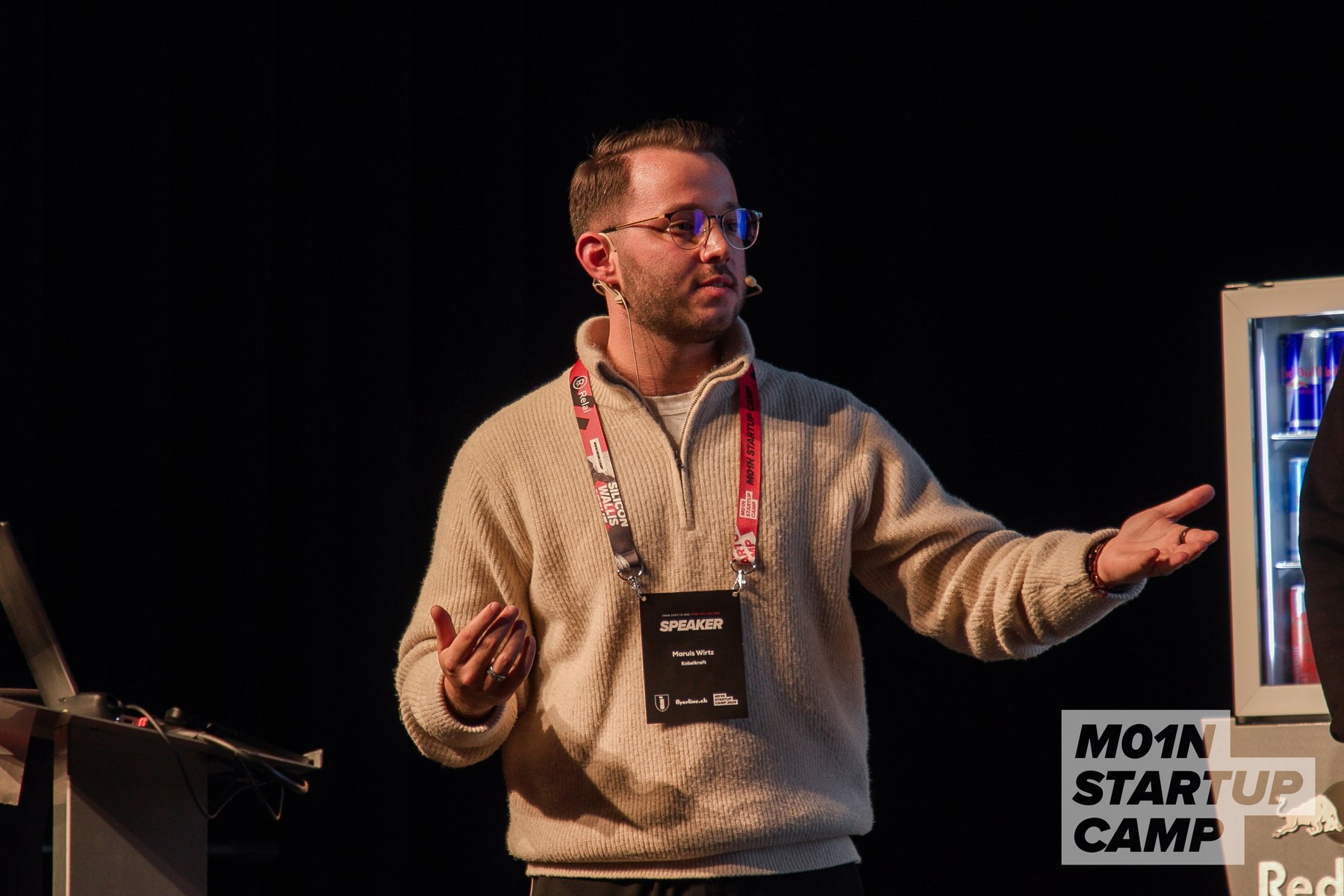 Marius Fuxs auf der Bühne des M01N Startup Camps, mit Brille und erklärender Handbewegung